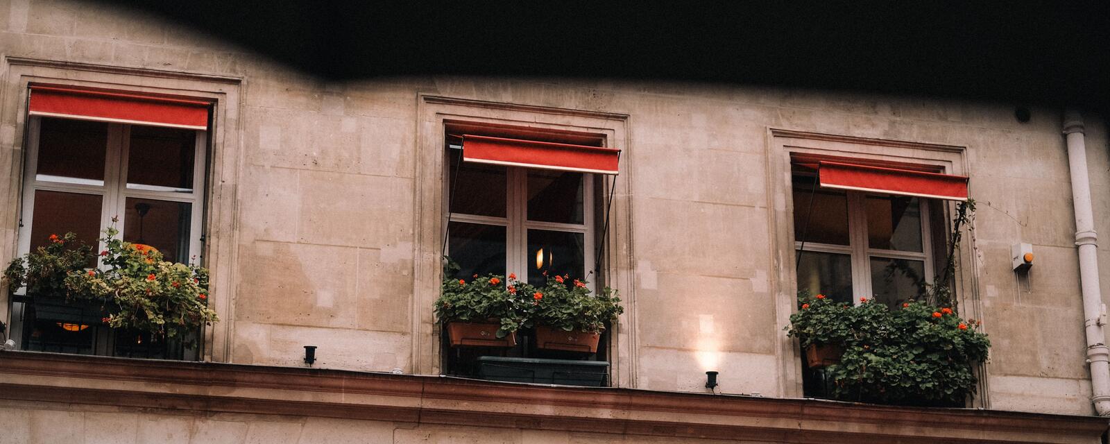 Three red awnings above flower-filled windows on a warm stone facade.